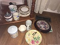 Photo showing a selection of four ceramic mugs, ceramic sugar container with lid, small cream pitcher, stacked decorative plates, and a decorative tray with floral design.