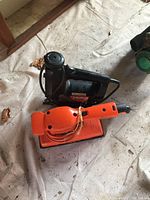 Black Model 31 variable speed scroll/saber saw and orange Black & Decker single action sander on floor with visible dust and cords.