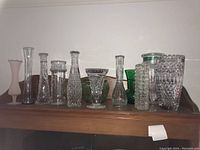 Photo of 12 assorted glass vases on wooden shelf showing variety in shape, pattern, and colors including clear, green, and pink frosted glass.