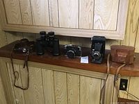 Overview of all items on wooden shelf: two camera cases, binoculars, three cameras.