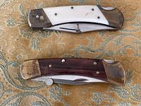 Top-down photo showing two vintage folding pocket knives placed parallel. One knife has a white composite handle with visible brass bolsters; the other knife has a darker wood handle with brass bolsters and rivets.