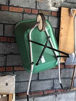Green metal wheelbarrow seen from the top and side showing wheel, handles and frame structure leaning against a brick wall.