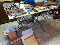 View of assembled Black & Decker Workmate 400 with a plywood top covered in various items. Bench is blue metal frame with attached plywood top and built-in vise.