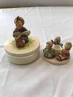 Image showing a round trinket dish with a seated boy playing guitar figurine and an angel-themed item with three children angels around a central holder, placed on a white cloth background.