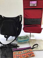 Front view of black Travelon purse showing front zippered pocket and adjustable strap, with assorted jewelry bags and red Reisenthal travel bag in the background.