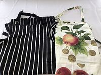 Two full-length aprons laid flat, one black with white vertical stripes and one white with botanical fruit print including peaches, showing front pockets and neck straps