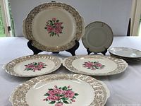 Four vintage Sebring platters with floral pink rose center and ornate 22k gold borders, shown on black stands and table