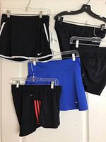 Five women's athletic skorts and shorts hanging on hangers, showing different colors and styles. Black Nike skort with white trim on the left, blue Nike skort in the center, black Adidas shorts with red stripes, and two more black bottoms.