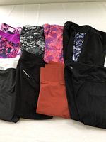 Top view of eight pairs of crop athletic leggings in various colors and patterns including black, rust, pinks, purples, and grayscale camo, folded and laid out on a white surface.