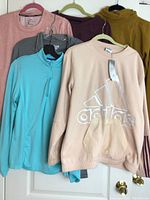 Six women's athletic long sleeve tops displayed on hangers, showing various colors and brands including Adidas, Peter Millar, Nike.