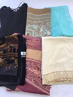 Photo showing six shawls and wraps neatly arranged, revealing various materials, colors, and patterns including silk, knit, paisley, velvet, and fringes.