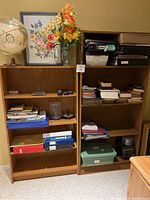 Photo showing both particleboard bookshelves side by side with items on their shelves, including books, binders, storage containers, and miscellaneous items.