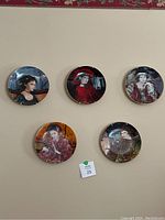 Five porcelain wall plates with portraits of women, arranged on wall, showing various characters in period attire.