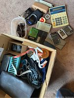 Photo showing assortment of calculators, sunglasses in cases, tape measures, a digital camera, and small accessories arranged on carpet.