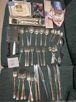 Overview of assorted mixed silver plate cutlery laid out on fabric surface, including forks, spoons, knives, and serving utensils.