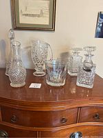 Six large glass pieces on wooden dresser: four cut-glass decanters with stoppers, one ice bucket, one pitcher. One stopper base damaged.