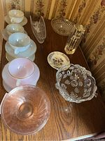 Overview of glassware including white Arcopal teacups and saucers, pink depression glass bowls, floral etched clear bowls, small floral dish, and rectangular Wedgwood ashtray.