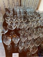Photo showing a large variety of glassware arranged on a table, including crystal wine glasses with cut patterns, champagne flutes, and other stemware.