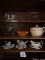 Full view of the shelves displaying all 8 items: two large clear etched glass bowls with floral and bird designs, one smaller brown ceramic bowl, a white ceramic tureen with lid, a white gravy boat with underplate, and three ceramic bird figurines placed on doilies.