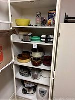 Photo of kitchen cupboard shelves containing various cookware, crockpots, bowls, and small appliances stacked together.