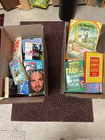 Two boxes of assorted books including visible titles such as 'The Sleeping Beauty's,' 'Classic Sports Quotes,' and fiction novels including Robert Harris's 'Enigma'.