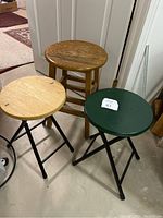 Three stools: one non-folding wooden stool with natural finish, two folding stools with black metal legs and wooden seats (light wood and dark green).