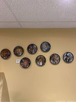 Photo showing 8 collector plates hanging on a wall in a horizontal line. Plates have various detailed images featuring romantic and medieval themes with blue patterned borders.