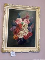 Full view of the floral oil painting in a carved and distressed white and gold wooden frame.