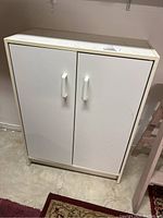 Front view of small white laminate cabinet with two doors and white handles, placed on a concrete floor against a wall.