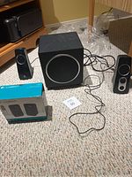 Photo showing a used Logitech speaker system including a subwoofer and two satellite speakers on carpet near table legs.