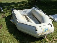 Top-side view of white inflatable dinghy with visible wear, two locking oars attached, and Baltic emblem on bow.
