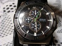 Front face of Guess men's watch showing black dial, subdials, and silver-tone bezel with minute markers; crystal shows scuffing.