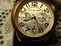 Close-up of watch face showing white dial with rose gold tone large Arabic numerals, three subdials, and date window.