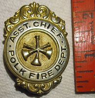 Close-up showing gold tone metal badge with ornate edges and three crossed bugles in center, text 'ASST. CHIEF POLK FIRE DEPT' around bugles.