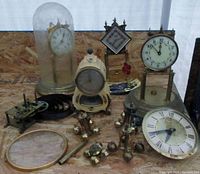Overall view of the lot showing multiple clocks, clock faces, glass covers, weights, and parts.