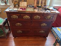 Front view of the antique chest of drawers with four drawers, marble top, and decorative brass handles. The chest shows wear on the wood and has various miscellaneous items placed on top.
