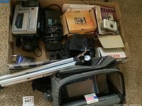 Overview of lot including cameras, tripod, binoculars, and storage box in cardboard.