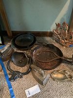 Photo showing two cast iron skillets, a large metal ice tong, one decorative metal bell, a sad iron with wooden handle, and a small decorative tray.