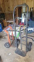 Two convertible hand trucks leaning against each other in a shed with visible wear and mechanical parts including frame, handles, wheels, and base plates.