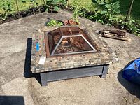 Full view of square mosaic tile fire pit located on concrete with garden visible in background.