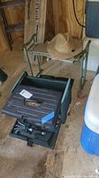 Green metal foldable gardening stool with fabric seat, tan woven gardening hat on top, alongside black and green wheeled gardening seat with storage and multiple gardening tools inside compartments.