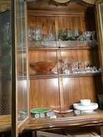 Shows a wooden cabinet shelf displaying various glassware items including clear glass stemware, a vintage amber bowl, and other glass bowls and plates