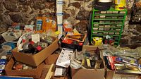 Wide view of boxes and items spread on workbench and floor including automotive headlights, electrical boxes, various hardware, and small tools in boxes and containers.