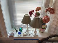 Two table lamps with beige shades, Mikasa vase with faux flowers, glass insulator, paperweight, small bells, and framed item arranged on windowsill.