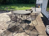 Full view of metal Woodard style patio set with round mesh table, 4 matching chairs, and outdoor cover on concrete patio.