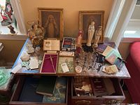 Wide shot displaying religious statues, framed art, rosaries, booklets, and books on a marble surface and shelf beneath.