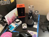 Multiple electronic items including JBL Flip 3 wireless speaker in box, Elite wireless speaker with box, activity tracker watch, Amazon Echo Dot speaker, and digital guitar tuner on a table.