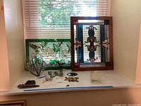 Two framed stained glass panels with floral and geometric designs on a window ledge, along with several small decorative glass ornaments including a wire heart, butterfly, and other glass art pieces.