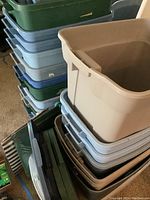 Stack of plastic storage bins in various colors including light blue, beige, dark gray, and green. Some lids are separate; boxes stacked on floor.