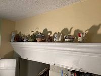 Wide view of mantel displaying 11 assorted pottery and figural tea pots and pitchers